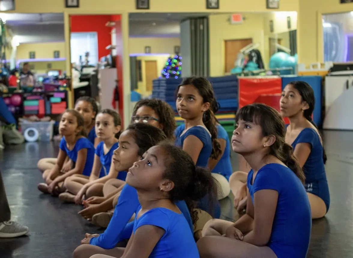 A row of young dancers seated on the floor listening closely to their teacher